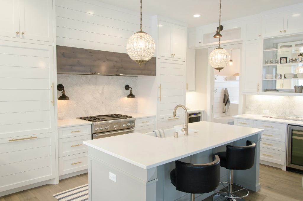 A modern white kitchen with a central island, two black bar stools, a stainless steel stove, marble backsplash, and two hanging pendant lights. The cabinets are white with gold handles.