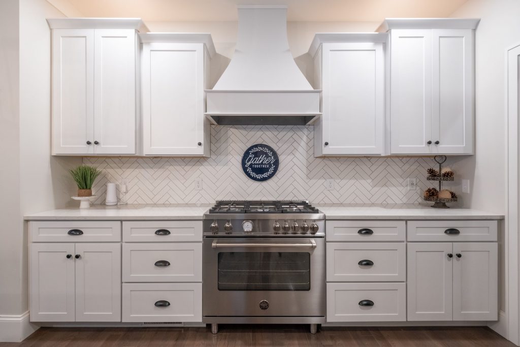 A modern kitchen features white cabinetry, a stainless steel oven with a hood, a herringbone-patterned backsplash, and a small plant and decorative items on the counter.